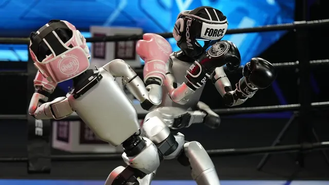 Two Unitree humanoid robots wearing boxing headgear and gloves, one pink and one black, squaring off inside a ring.