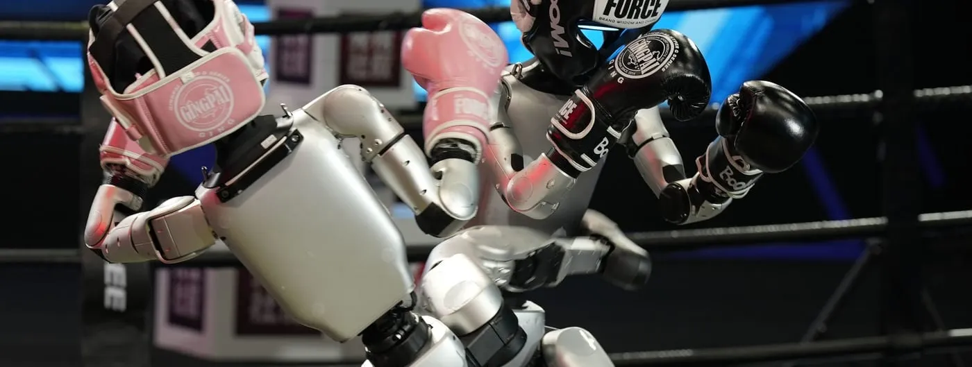 Two Unitree humanoid robots wearing boxing headgear and gloves, one pink and one black, squaring off inside a ring.