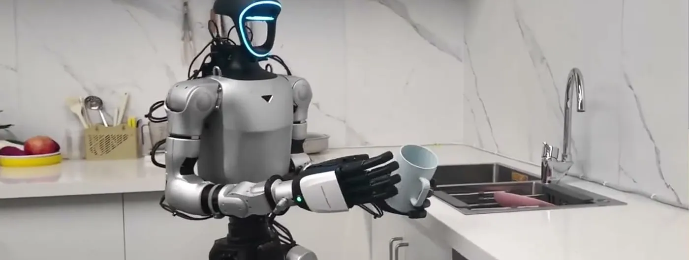 A Unitree humanoid robot with a glowing blue visor stands at a kitchen counter, gripping a light-blue mug with its five-finger hand beside a fruit bowl and a utensil holder.