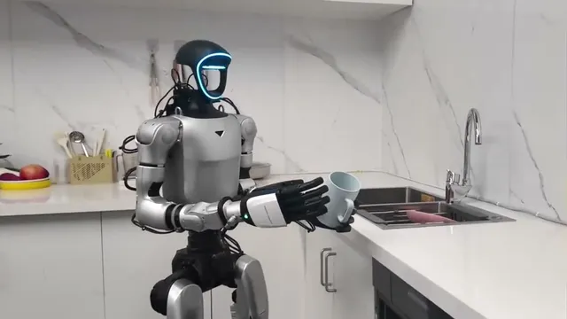 A Unitree humanoid robot with a glowing blue visor stands at a kitchen counter, gripping a light-blue mug with its five-finger hand beside a fruit bowl and a utensil holder.