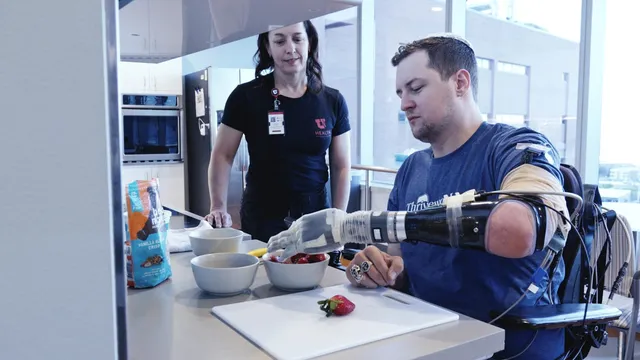 A trial participant uses a prosthetic LUKE bionic arm wired to sensors on his residual limb to pick up a strawberry from a bowl at a kitchen counter while a clinician looks on.