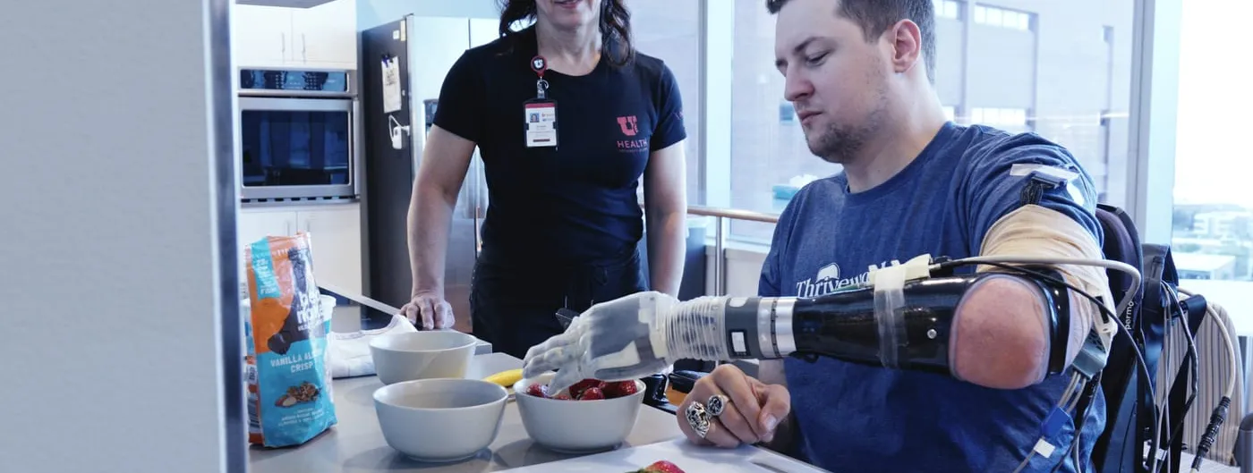 A trial participant uses a prosthetic LUKE bionic arm wired to sensors on his residual limb to pick up a strawberry from a bowl at a kitchen counter while a clinician looks on.
