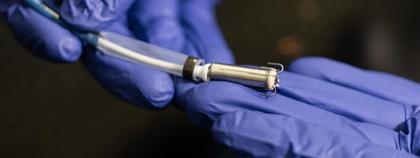 Two blue-gloved hands hold a small metal cylinder about the size of a fingertip, with tines at one end and a white sheath and thin cable at the other.