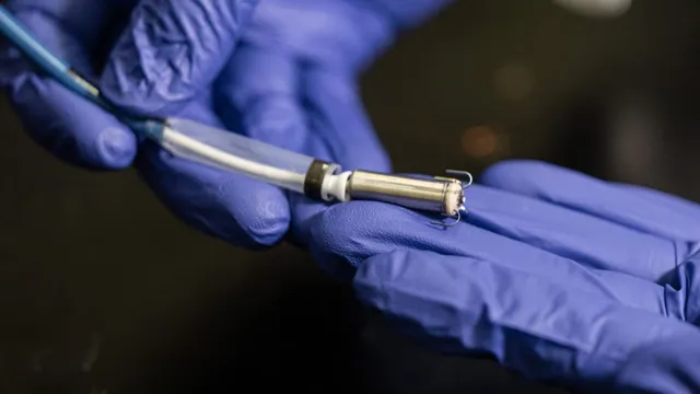 Two blue-gloved hands hold a small metal cylinder about the size of a fingertip, with tines at one end and a white sheath and thin cable at the other.