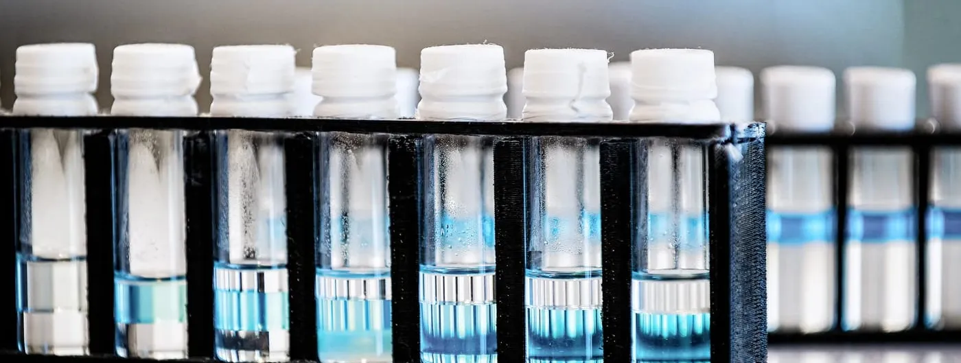 Close-up of a black rack of capped laboratory vials holding clear and blue liquid on a bench in a research laboratory, with a second rack blurred in the background.