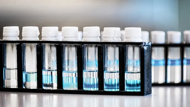 Close-up of a black rack of capped laboratory vials holding clear and blue liquid on a bench in a research laboratory, with a second rack blurred in the background.