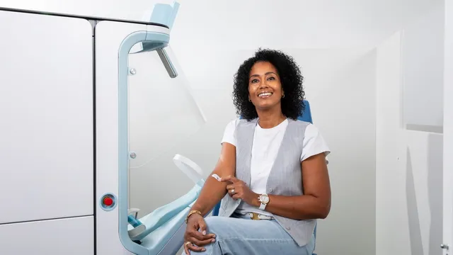 A woman sits in a blue clinic chair beside the white Aletta cabinet, a bandage on her inner arm, with the machine and its padded arm cradle to her left.