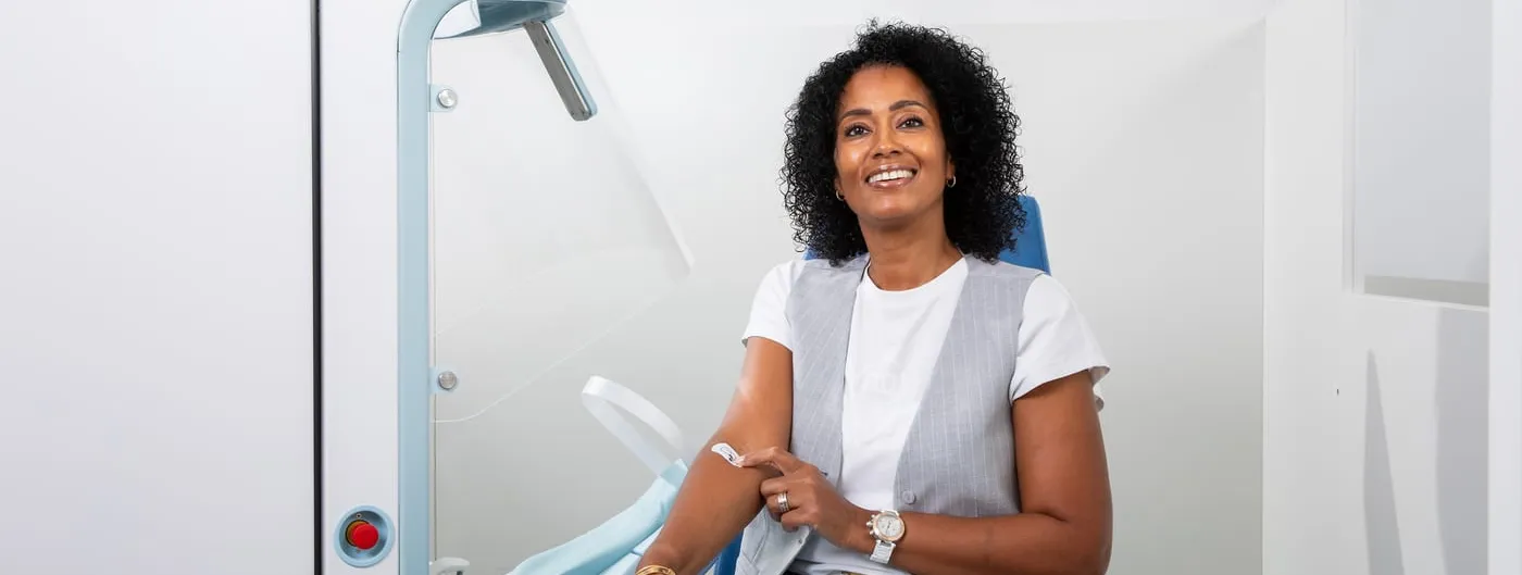A woman sits in a blue clinic chair beside the white Aletta cabinet, a bandage on her inner arm, with the machine and its padded arm cradle to her left.