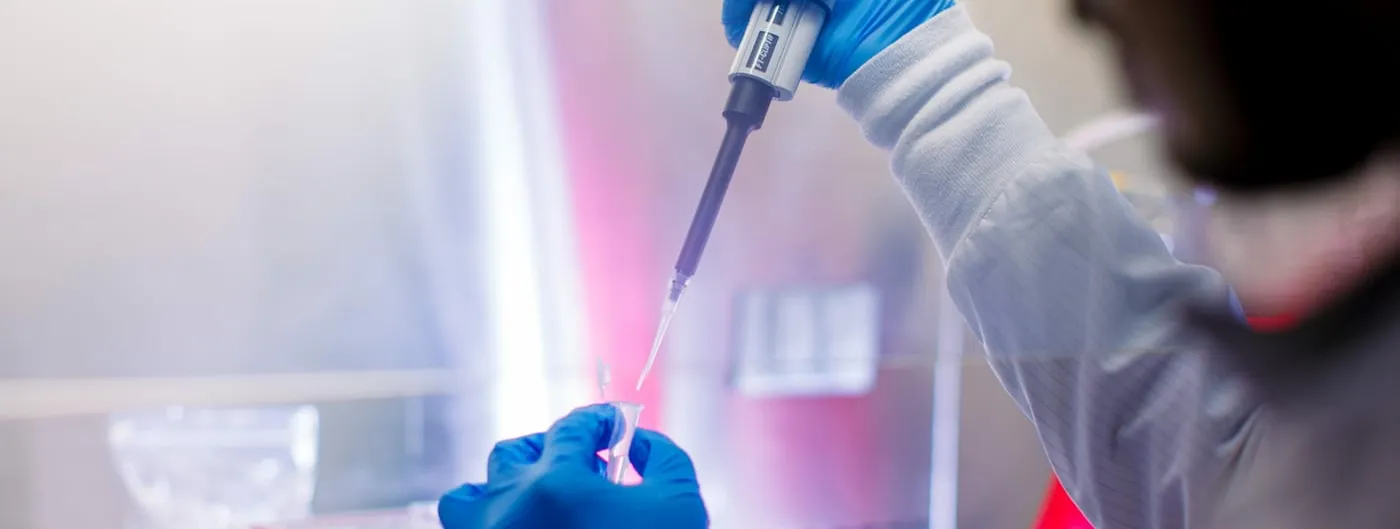 A gloved researcher pipettes a sample into a small vial inside a lit laboratory biosafety cabinet.