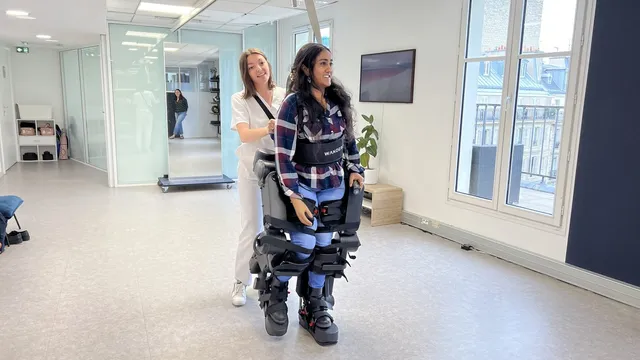 A woman stands upright inside the black Atalante X exoskeleton, its rigid leg segments strapped from boot to hip and a torso brace at her waist, while a clinician in white steadies her from behind in a bright clinic room.