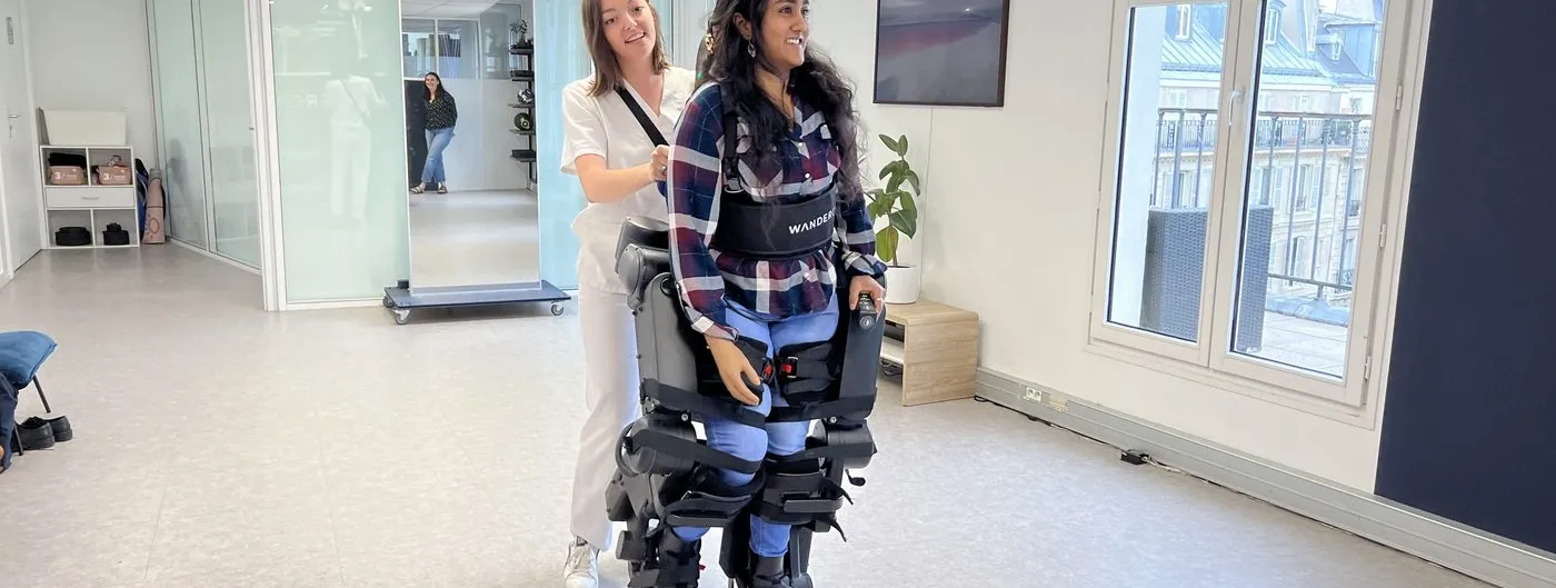 A woman stands upright inside the black Atalante X exoskeleton, its rigid leg segments strapped from boot to hip and a torso brace at her waist, while a clinician in white steadies her from behind in a bright clinic room.