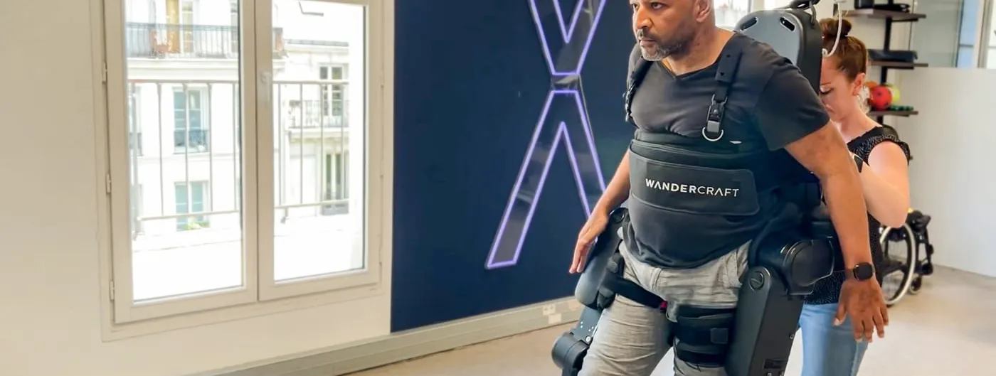 A man wearing a Wandercraft Atalante X exoskeleton walks upright in a rehabilitation room, with a therapist steadying the device from behind and a large illuminated X on the wall.
