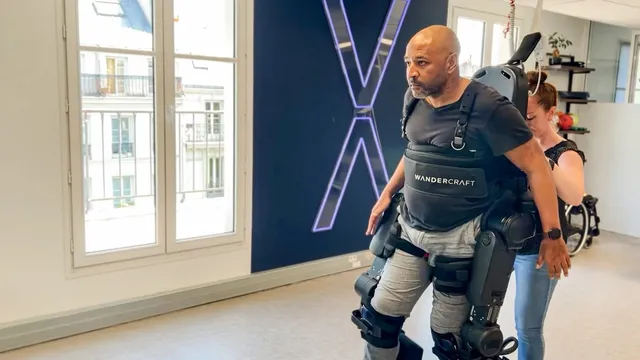 A man wearing a Wandercraft Atalante X exoskeleton walks upright in a rehabilitation room, with a therapist steadying the device from behind and a large illuminated X on the wall.