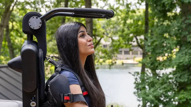 A woman with long dark hair stands upright on a wooden deck beside a lake, held by a black exoskeleton frame that runs up her back with padded shoulder straps and a red-buckled chest harness.