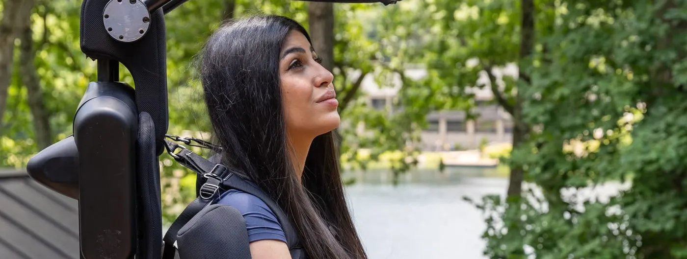 A woman with long dark hair stands upright on a wooden deck beside a lake, held by a black exoskeleton frame that runs up her back with padded shoulder straps and a red-buckled chest harness.