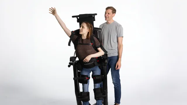 A woman stands upright in the Eve exoskeleton with one arm raised, a companion beside her, photographed against a plain studio backdrop.