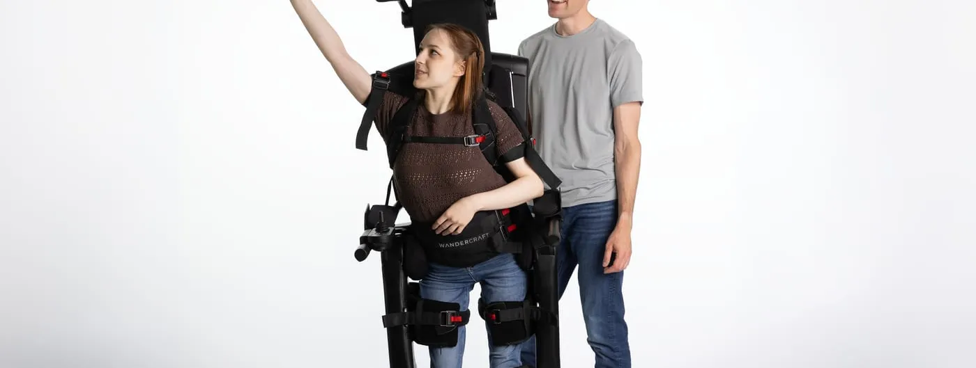 A woman stands upright in the Eve exoskeleton with one arm raised, a companion beside her, photographed against a plain studio backdrop.