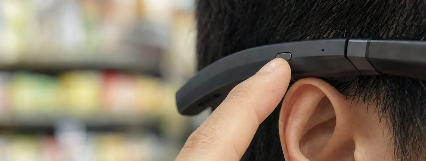 A close side view of dark-framed smart glasses worn on a head, with a fingertip pressing the temple arm just in front of the ear and blurred shop shelving behind.