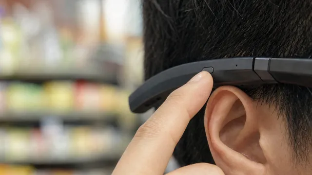 A close side view of dark-framed smart glasses worn on a head, with a fingertip pressing the temple arm just in front of the ear and blurred shop shelving behind.
