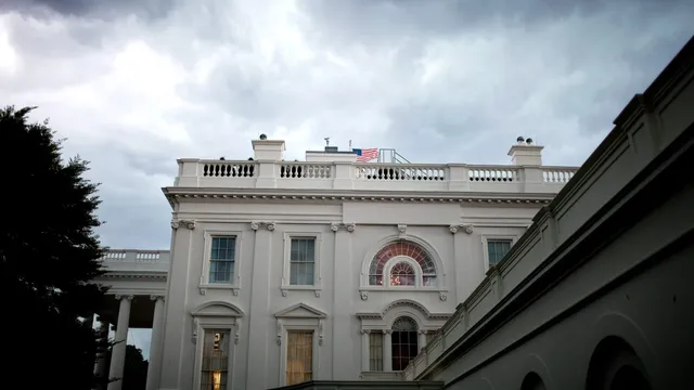 The upper floors of the White House's west side under heavy grey storm clouds, with an American flag flying above the balustrade and a dark tree in the left foreground.