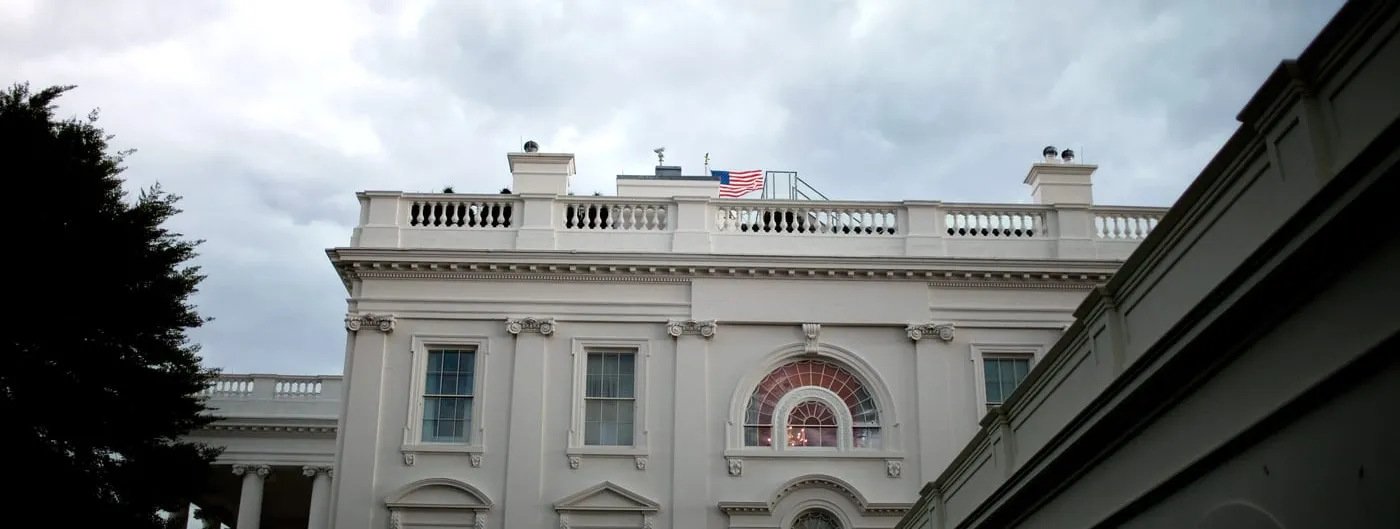 The upper floors of the White House's west side under heavy grey storm clouds, with an American flag flying above the balustrade and a dark tree in the left foreground.