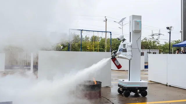 A white humanoid robot mounted on a wheeled column aims a red fire extinguisher at a burning steel drum on wet pavement, its spray crossing a yard filled with smoke.