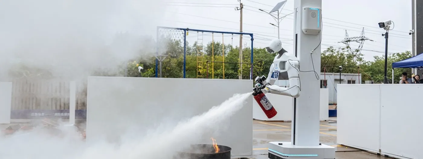 A white humanoid robot mounted on a wheeled column aims a red fire extinguisher at a burning steel drum on wet pavement, its spray crossing a yard filled with smoke.