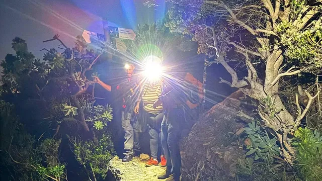 At night on Table Mountain's rocky Platteklip Gorge trail, two Wilderness Search and Rescue members flank an exhausted hiker wearing a hip-and-thigh exoskeleton, lit by a bright headlamp.