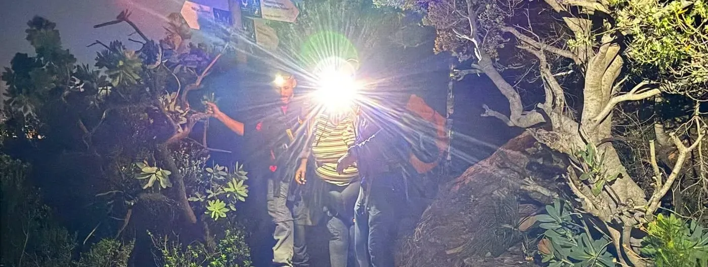 At night on Table Mountain's rocky Platteklip Gorge trail, two Wilderness Search and Rescue members flank an exhausted hiker wearing a hip-and-thigh exoskeleton, lit by a bright headlamp.