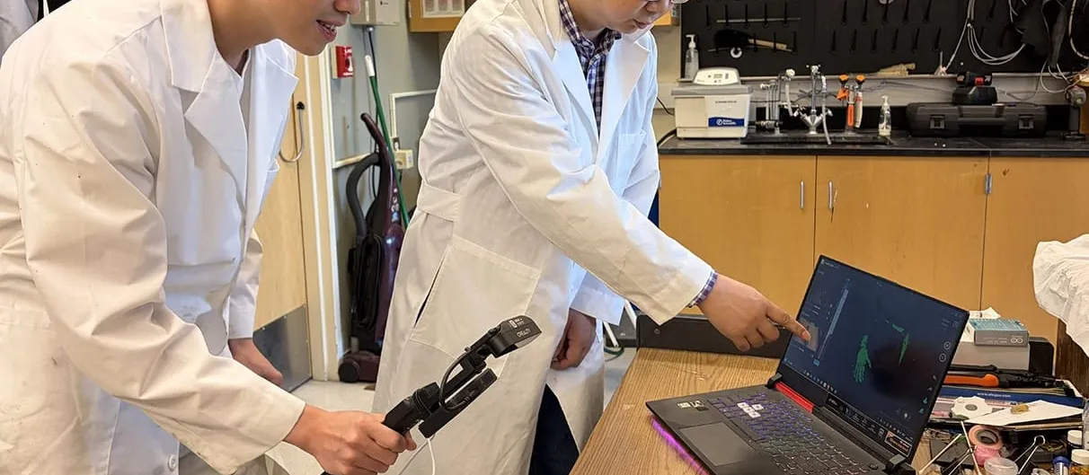 Two researchers in white lab coats at a cluttered workbench; one holds a black handheld sensor probe while the other points at a laptop screen showing a green sensor readout.