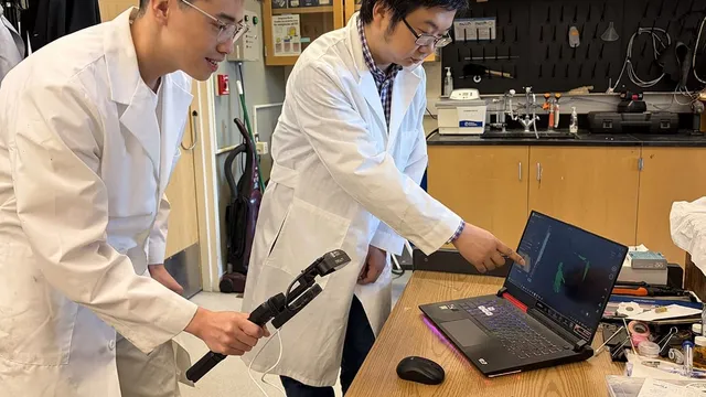 Two researchers in white lab coats at a cluttered workbench; one holds a black handheld sensor probe while the other points at a laptop screen showing a green sensor readout.
