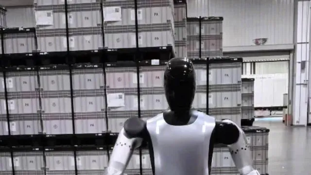 A bipedal humanoid robot with a glossy black head and white-and-black torso, seen from behind, standing in a factory warehouse in front of stacked grey storage crates.