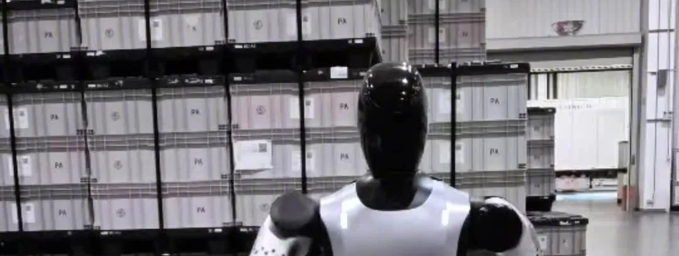 A bipedal humanoid robot with a glossy black head and white-and-black torso, seen from behind, standing in a factory warehouse in front of stacked grey storage crates.