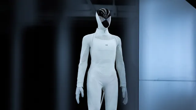 A white full-size humanoid robot in a knitted body covering with a dark reflective faceplate, standing in a dimly lit industrial hall.