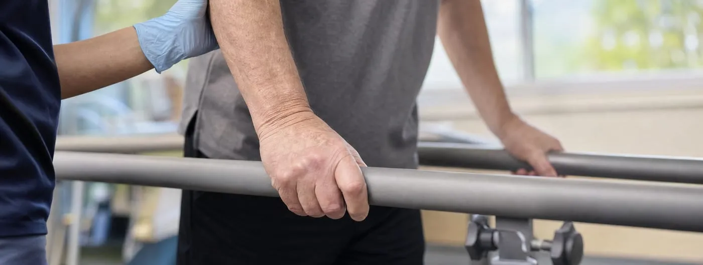 An older adult grips a padded parallel bar with both hands during a supervised walking test while a clinician in blue steadies their forearm with a gloved hand.