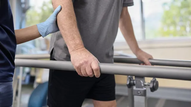 An older adult grips a padded parallel bar with both hands during a supervised walking test while a clinician in blue steadies their forearm with a gloved hand.