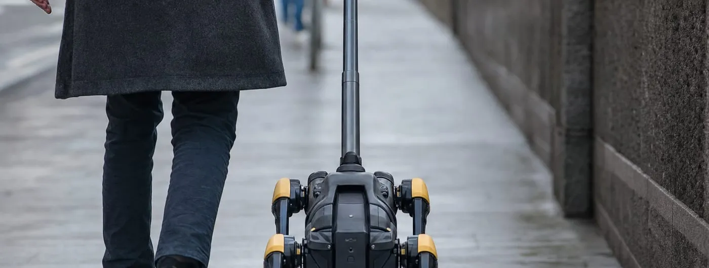 A four-legged robot with wheels on its feet and a tall rigid handle moves along a wet city pavement beside a walking person seen from behind.