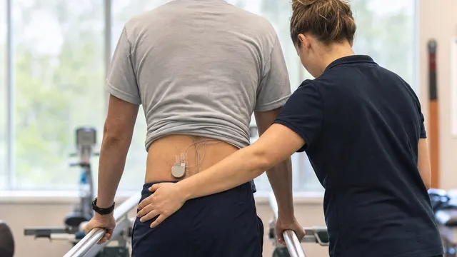 Seen from behind, a patient stands between parallel bars in a rehabilitation gym, a small implanted pulse generator and thin leads visible at the lower back, while a therapist steadies them at the hip.