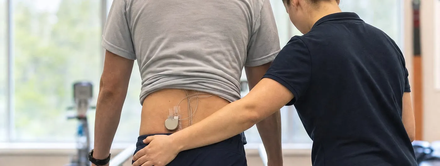Seen from behind, a patient stands between parallel bars in a rehabilitation gym, a small implanted pulse generator and thin leads visible at the lower back, while a therapist steadies them at the hip.